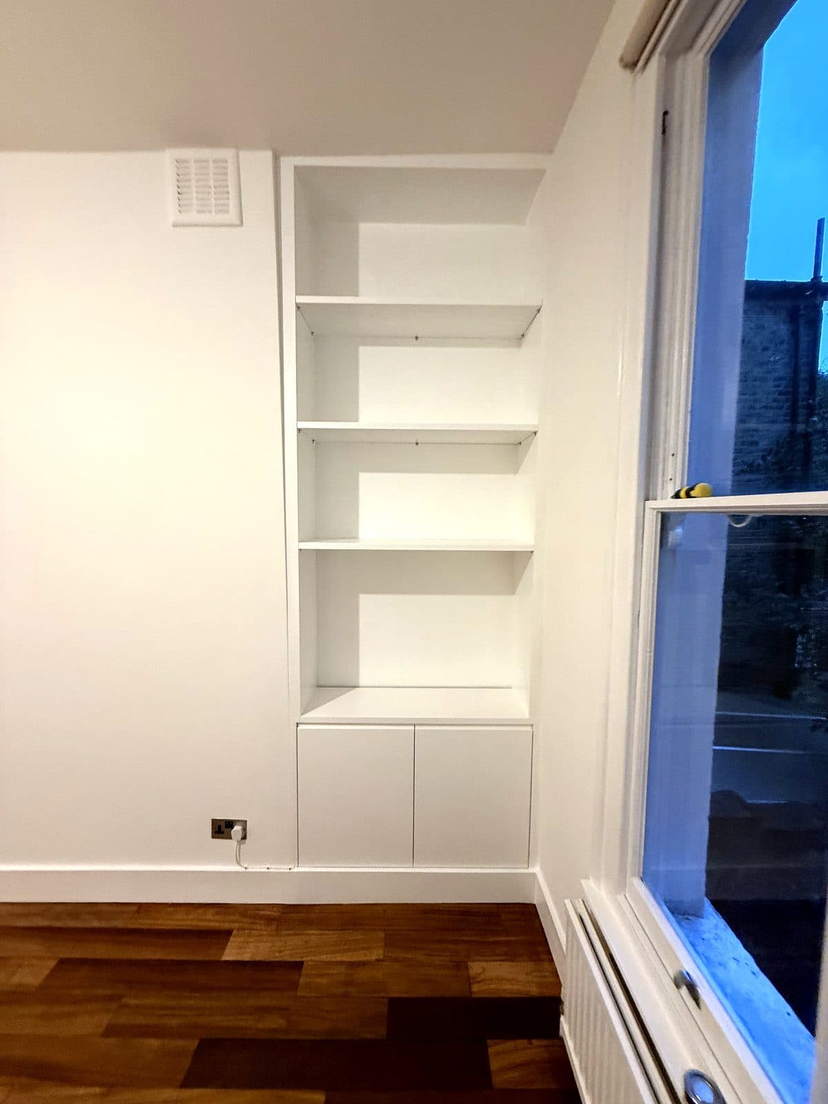 White painted alcove unit with open shelves above a low two-door cupboard, next to a sash window