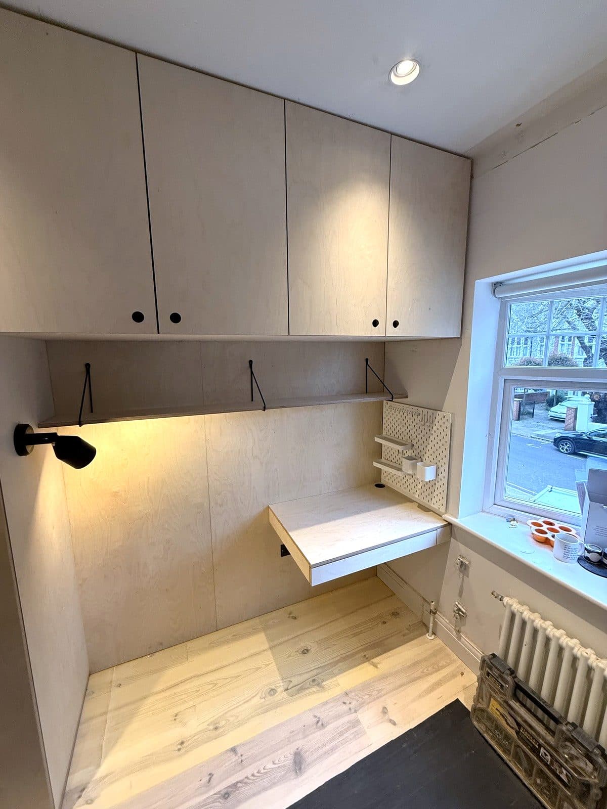 Birch ply room with a tall wardrobe, overhead cupboards and a wall-hung desk lit by a wall lamp